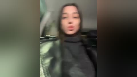 Media: Blurry video of a woman with light skin, brown hair, and wearing a black turtleneck sweater, partially visible, in a dimly lit room with indistinct background objects.