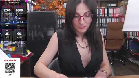 Media: Video of a woman with long dark hair, wearing red-framed glasses, a black sleeveless dress, and a silver necklace, seated at a desk in an office.