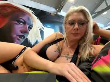 Media: Video of a fair-skinned, blonde woman in glasses and black lingerie, lying on a table, with a large, colorful, abstract mural in the background.