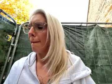 Media: Video of a middle-aged blonde woman with glasses, wearing a white hoodie, standing outdoors near a green tarp and a building, with autumn leaves in the background.