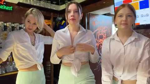 Media: Video of three women in white button-up shirts and light green skirts, standing in a dimly lit bar with neon signs and shelves of glasses in the background.