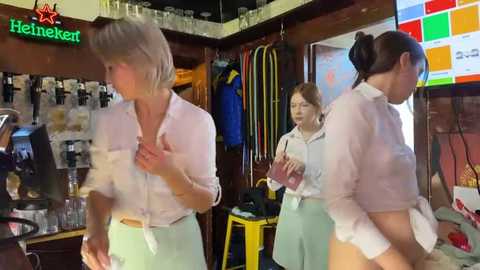 Media: Video of three women in a cozy, dimly-lit bar. One woman in a pink shirt and green skirt is dancing; another in a white top and green pants watches; a third in a white top and green pants holds a glass.