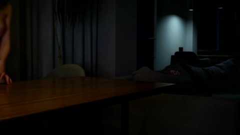 Media: A dimly lit video of a person lying on a couch under a low ceiling, with a wooden table in the foreground, creating a moody, intimate atmosphere.