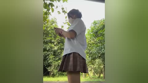 Media: Video of a person in a white shirt and brown plaid skirt, holding a small, white object, standing outdoors among lush greenery.