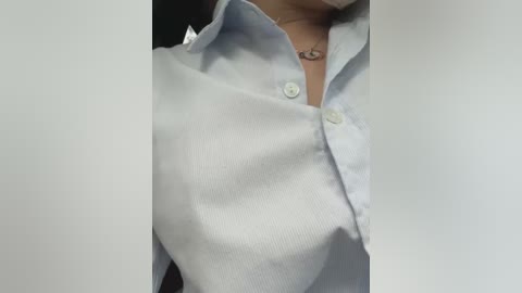 Media: Video of a partially unbuttoned light blue shirt with a white collar, revealing a necklace and part of the chest. The background is out of focus, emphasizing the fabric texture.