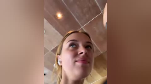 Media: Video of a young woman with fair skin, light blonde hair, and blue eyes, smiling, looking upward. She's wearing white earbuds. Background features a beige tiled ceiling and a light bulb.