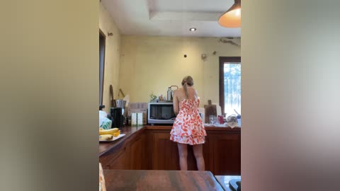 Media: Video of a woman with blonde hair, wearing a floral dress, cooking in a cozy, rustic kitchen with wooden cabinets and yellow walls.