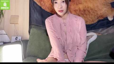 Media: A video shows an East Asian woman with light skin, brown hair, and red lipstick, wearing a pink ruffled blouse, sitting on a green couch, surrounded by a cozy, eclectic room with a large, abstract wall hanging.