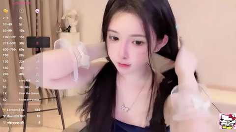 Media: Video of a fair-skinned, slender Asian woman with long black hair, adjusting her hair in a bedroom. She wears a dark top and necklace, with a digital overlay displaying viewer count and social media icons.