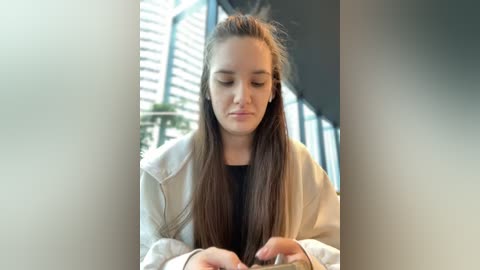 Media: Video of a young woman with long, straight brown hair, wearing a white jacket, seated indoors, focused on her smartphone, blurred background with vertical window blinds.