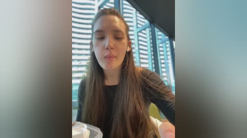 Media: A video of a young woman with long, straight brown hair, wearing a black long-sleeve shirt, seated indoors. She has her eyes closed and appears to be deep in thought. The background features vertical blinds and a modern, minimalist interior.