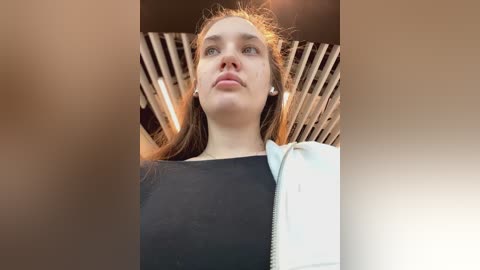 Media: Video of a young woman with fair skin and brown hair, wearing a black top, standing in a modern, light-filled room with slatted wooden ceiling panels. She holds a white bag in her right hand.