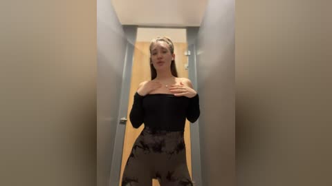 Media: A video of a slim, fair-skinned woman with long blonde hair, wearing a black off-the-shoulder top and grey camo pants, standing in a narrow hallway.