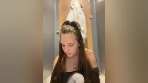 Media: Video of a young woman with long brown hair in a ponytail, wearing a black top, sitting in a locker room with beige lockers and a white towel hanging behind her.