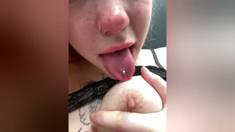 Media: A close-up video of a woman with pale skin, pierced nose, and tongue piercing, licking her own pierced nipple, wearing black lace lingerie.