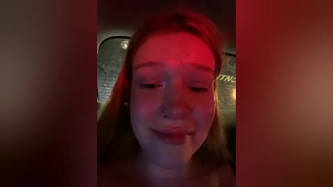 Media: A dimly lit video of a young woman with long, straight brown hair, wearing a nose ring, sitting in a car, with red lighting casting a shadow on her face.