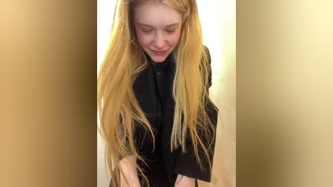 Media: Video of a young woman with long, straight, platinum blonde hair, wearing a black dress, standing against a plain beige wall.