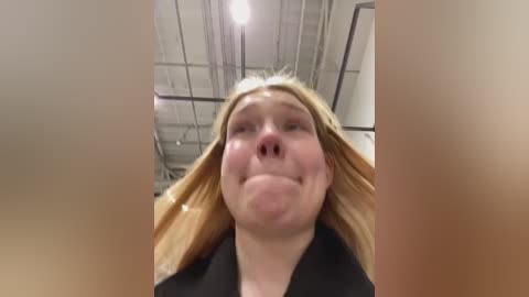 Media: Video of a blonde woman with long hair, wearing a black coat, making a silly face with her mouth open. Background shows a dimly lit, industrial room with exposed pipes and a low ceiling.