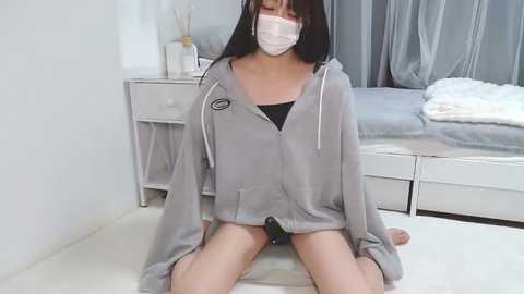 Media: Video of a young Asian woman with long black hair, wearing a gray hoodie, black panties, white mask, and seated on a light-colored floor in a minimalist bedroom with a bed and white dresser.