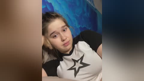 Media: Video of a young woman with fair skin, brown hair, and a neutral expression, wearing a black and white t-shirt with a star design, lying on a bed with a blue and white striped comforter.