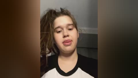 Media: Video of a young, light-skinned girl with tousled brown hair, wearing a black and white T-shirt, making a pouty face, standing in front of a blurry background.