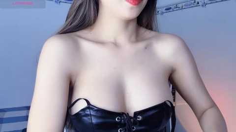 Media: Video of a fair-skinned woman with long brown hair, wearing a black, shiny, strapless corset with laces, partially revealing her large breasts, against a white background with a hint of blue and red lighting.