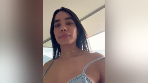 Media: A video of a young woman with long black hair and light skin, wearing a light blue spaghetti-strap tank top, standing in a bright, indoor setting with white walls and a ceiling.