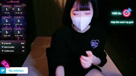 Media: Video of an East Asian woman with a mask, wearing a black shirt with a heart graphic, sitting on a bed, in a dimly lit room.