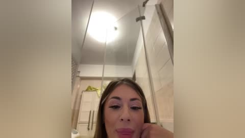 Media: A video of a woman with light skin and straight brown hair, wearing pink lipstick, taken from a low angle, looking up at a ceiling light. The bathroom background features a shower and beige tiles.