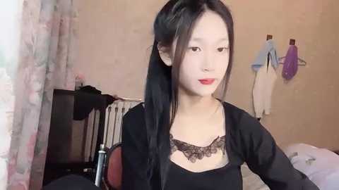 Media: Video of an East Asian woman with long black hair, red lipstick, and black lace top, sitting in a cluttered room with floral curtains, hanging clothes, and a bed in the background.