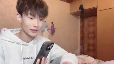 Media: Video of an Asian man with short dark hair in a white hoodie, holding a smartphone, in a dimly lit room with beige walls and a brick wall, featuring a person's silhouette and a purple towel hanging.