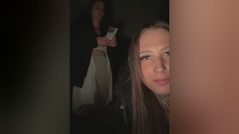 Media: Video of a blonde woman with fair skin, wearing a black jacket, holding a white phone, partially obscured by a dark vertical object. Background shows a dark room with another woman, partially visible, holding a white phone.