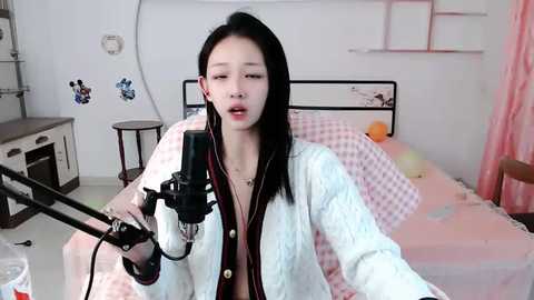 Media: Video of an Asian woman with long black hair singing into a microphone in a pink, patterned bedroom.