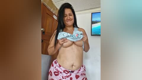 Media: Video of a smiling Asian woman with long black hair, topless, holding a light blue shirt, wearing pink pajama pants with red heart prints. Background shows a wooden door, wall, and a TV with a beach scene.