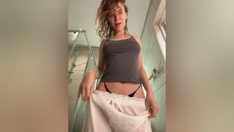 Media: Video of a young woman with wet, curly hair, wearing a gray tank top and black thong, lifting a white towel in a bathroom with tiled walls and a shower.