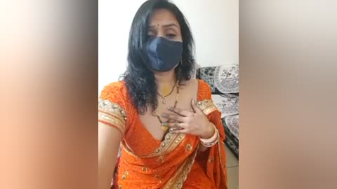 Media: Video of a woman in an orange sari with gold embroidery, wearing a black face mask, with her hands over her chest, in a room with patterned furniture and a neutral-colored wall.