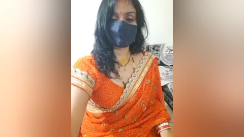 Media: Video of a woman with medium skin tone, dark hair, wearing an orange saree adorned with intricate gold embroidery, a black mask, and a yellow necklace, seated indoors.