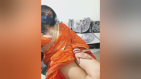 Media: Video of a South Asian woman with medium skin tone, wearing an orange sari with gold embroidery, sitting on a bed. Background includes a white wall, decorative black and white tapestry, and a blue mask covering her mouth.