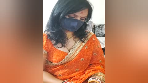 Media: Video of a woman in a vibrant orange saree with intricate gold embroidery, wearing a black face mask, and red dot on her forehead, seated in a home setting with blurred background.