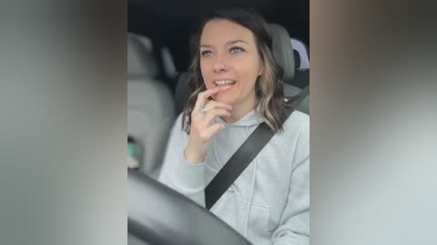 Media: Video of a woman with shoulder-length brown hair, wearing a light grey sweater, driving a car, holding her finger to her mouth in a playful manner, with a blurred background.