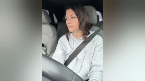 Media: Video of a woman in a car, wearing a light grey hoodie, with a grey seatbelt across her chest. She is looking straight ahead, seated in the driver's seat.