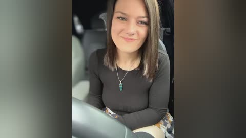 Media: A video of a young woman with straight, shoulder-length brown hair, smiling, wearing a black long-sleeve top and a green necklace. She sits in a car, partially framed by the window.