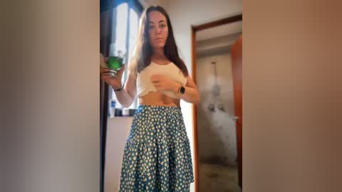 Media: Video of a young woman with long brown hair, wearing a white crop top and blue floral skirt, standing in a doorway, holding a green bottle.