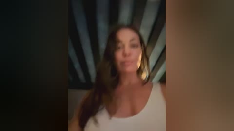 Media: A blurred video of a woman with long brown hair, wearing a white tank top, against a dark, dimly lit background with vertical stripes. Her facial expression is unclear.