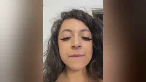 Media: A close-up video of a light-skinned woman with curly dark hair, wearing dramatic eyeliner and nude lipstick, looking serene with eyes closed.