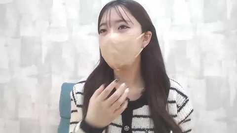 Media: A video of an East Asian woman with long black hair, wearing a black-and-white striped sweater, a surgical mask, and a blue bag, looking worried against a white, textured background.