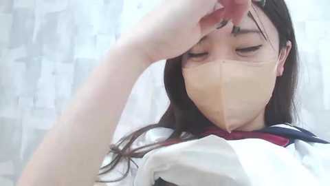 Media: A close-up video of an East Asian woman with long, dark hair, wearing a white and red sailor-style school uniform and a face mask, holding her hair away from her face, looking tired.