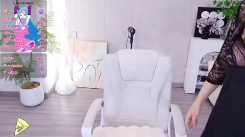 Media: Video of a modern, minimalist salon featuring a white massage chair, a white wall with a poster, a fern plant, and a person in a black lace dress.