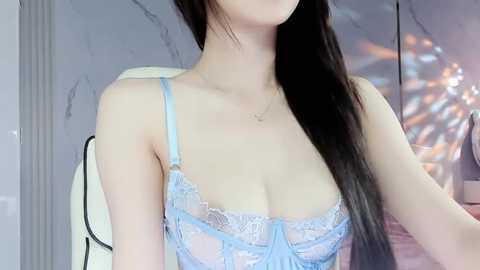 Media: Video of an Asian woman with long, straight black hair, wearing a delicate, light blue lace bra that reveals her ample breasts. She sits in a modern, minimalist room with a marble wall and a white chair.