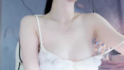 Media: Video of a slender, fair-skinned woman with long dark hair, wearing a white lace camisole, partially pulled down to reveal her cleavage. She has a small, delicate necklace on. Background features a marbled, light blue wall.